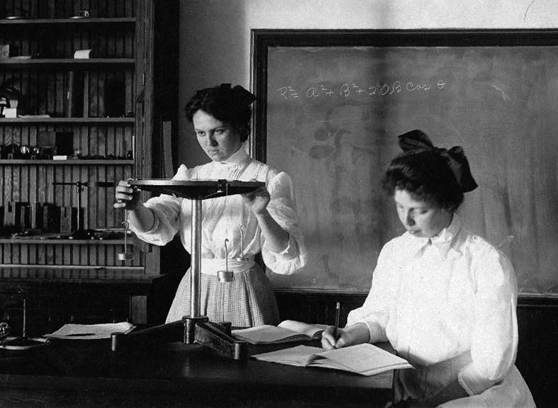 Two women in Edwardian-era dress use a scientific instrument and take notes