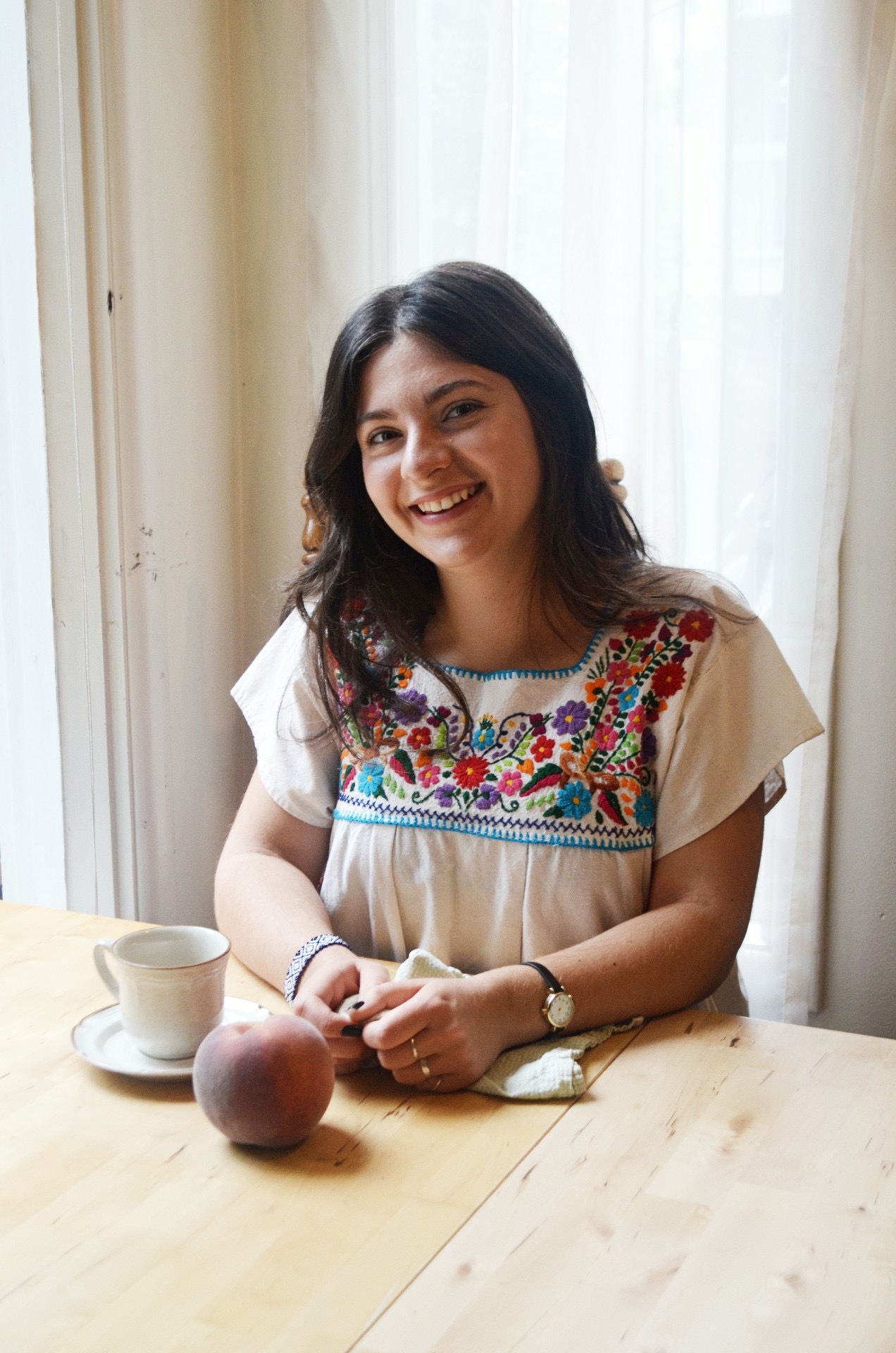 Serafima wearing an embroidered top sitting in soft natural light at a table with a coffee cup and a peach