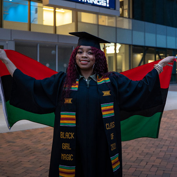 Jade in graduation cap and gown, holding a pan African flag behind her and wearing a green/yellow/red striped sash that reads, "Black Girl Magic, Class of 2021"